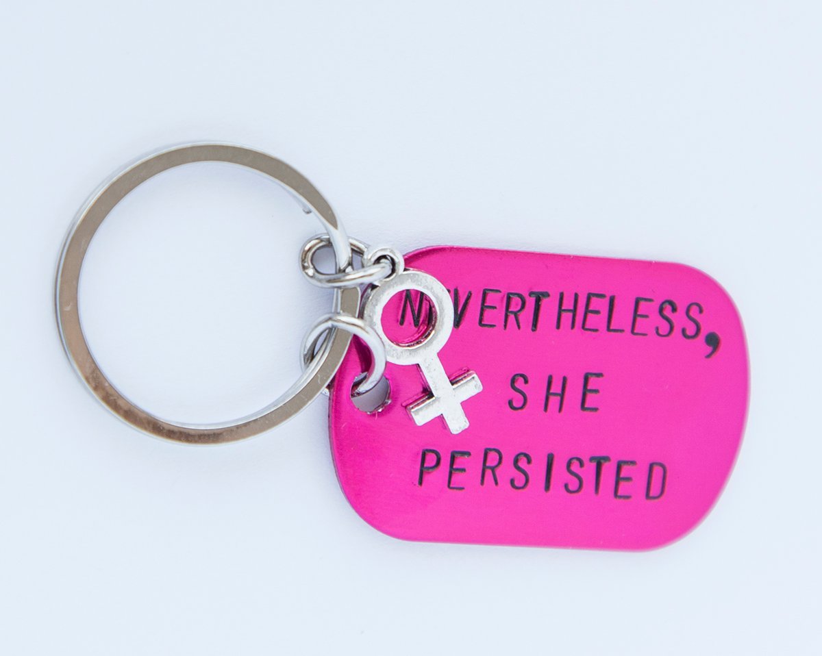 A pink feminist keychain with the phrase 'Nevertheless She Persisted' hand-stamped on a lightweight aluminum tag, attached to a durable stainless steel ring.